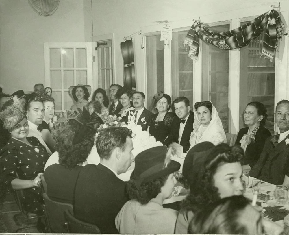 A black and white photo of people dressed formally, seated at a long table during a wedding celebration.