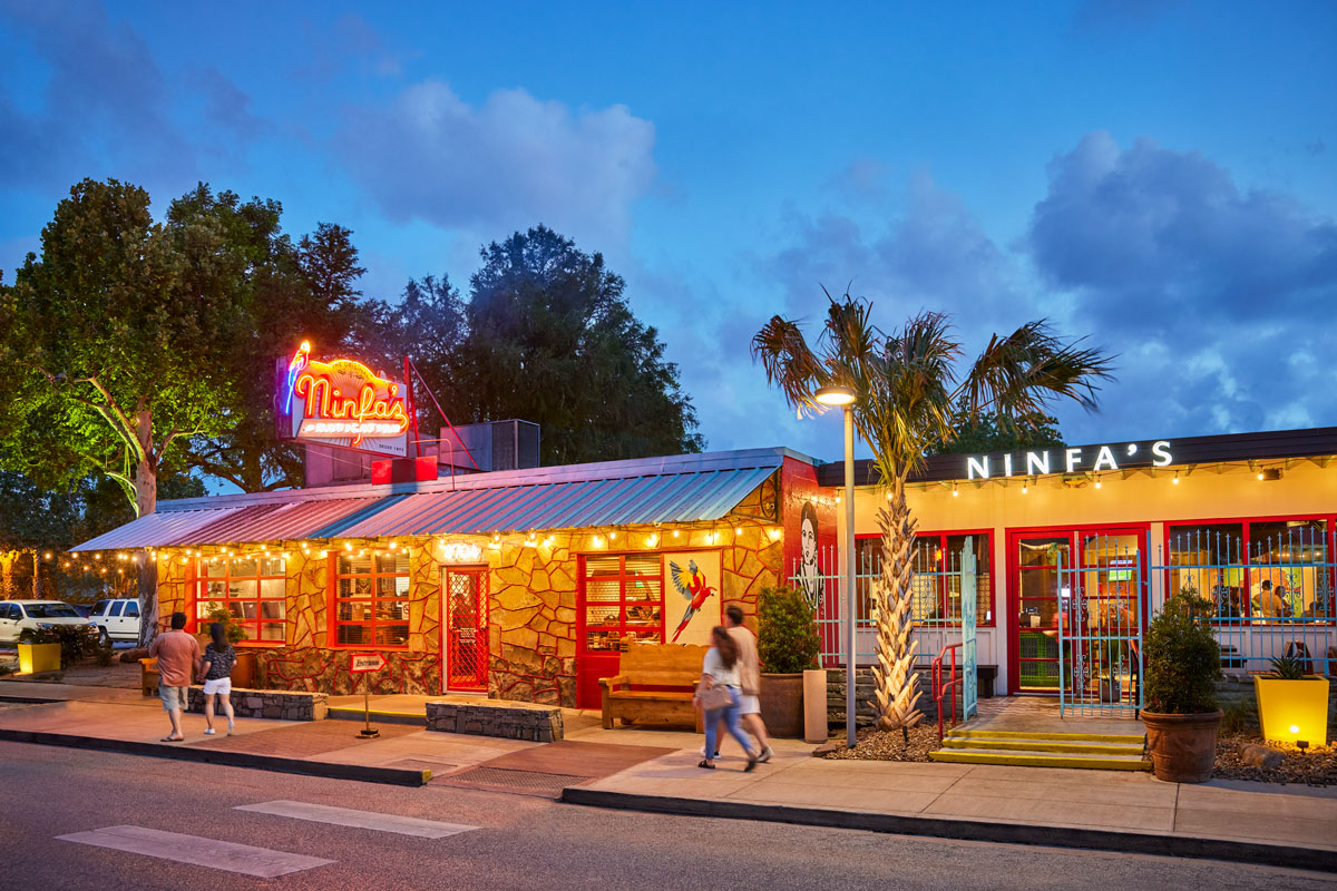 Colorful restaurant with neon Ninfas sign, outdoor lights, palm tree, and people walking nearby at dusk.