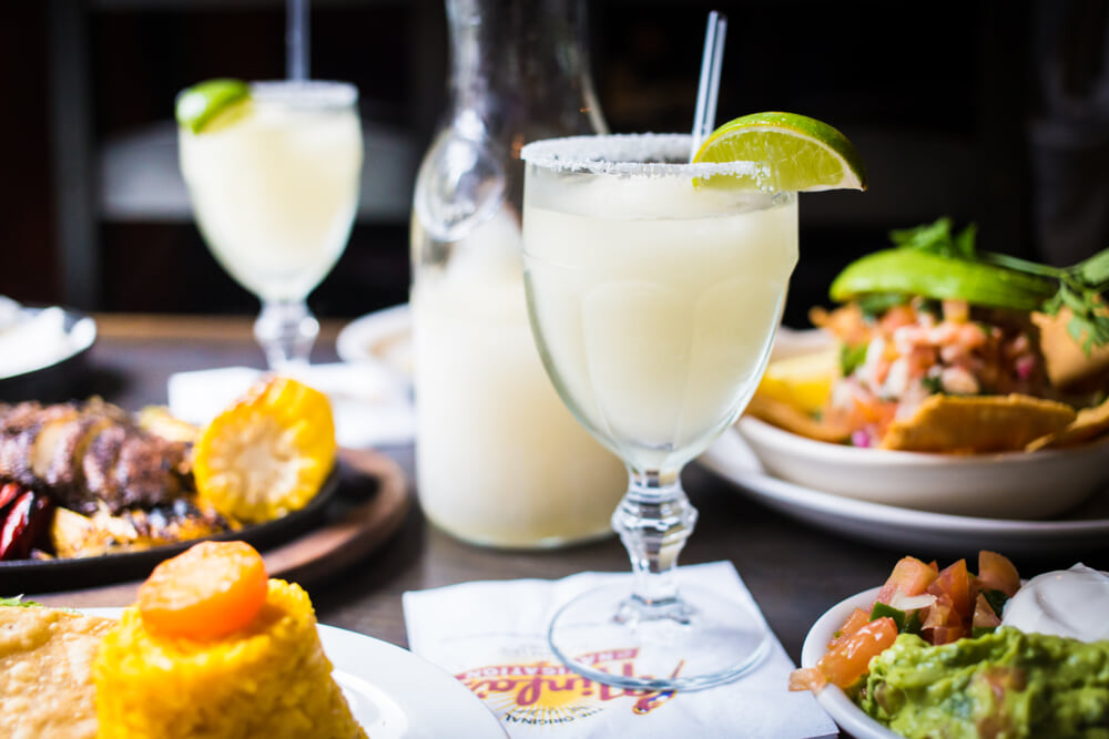 A Ninfa's margarita with a lime wedge on a table with Mexican food and another drink in the background.