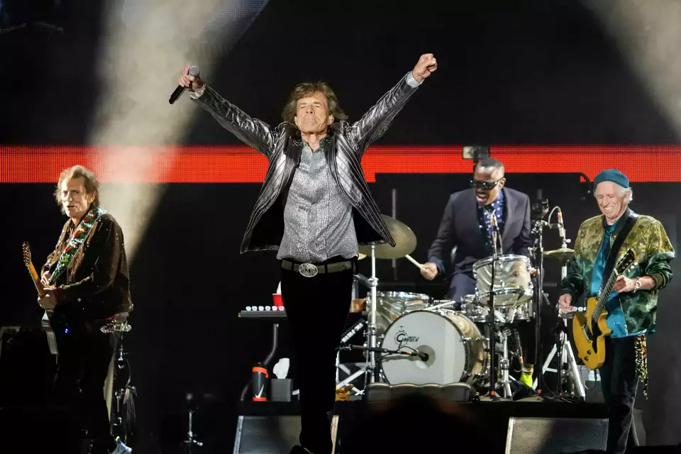 The Rolling Stones perform on stage under bright lights, with Mick Jagger raising both arms triumphantly.