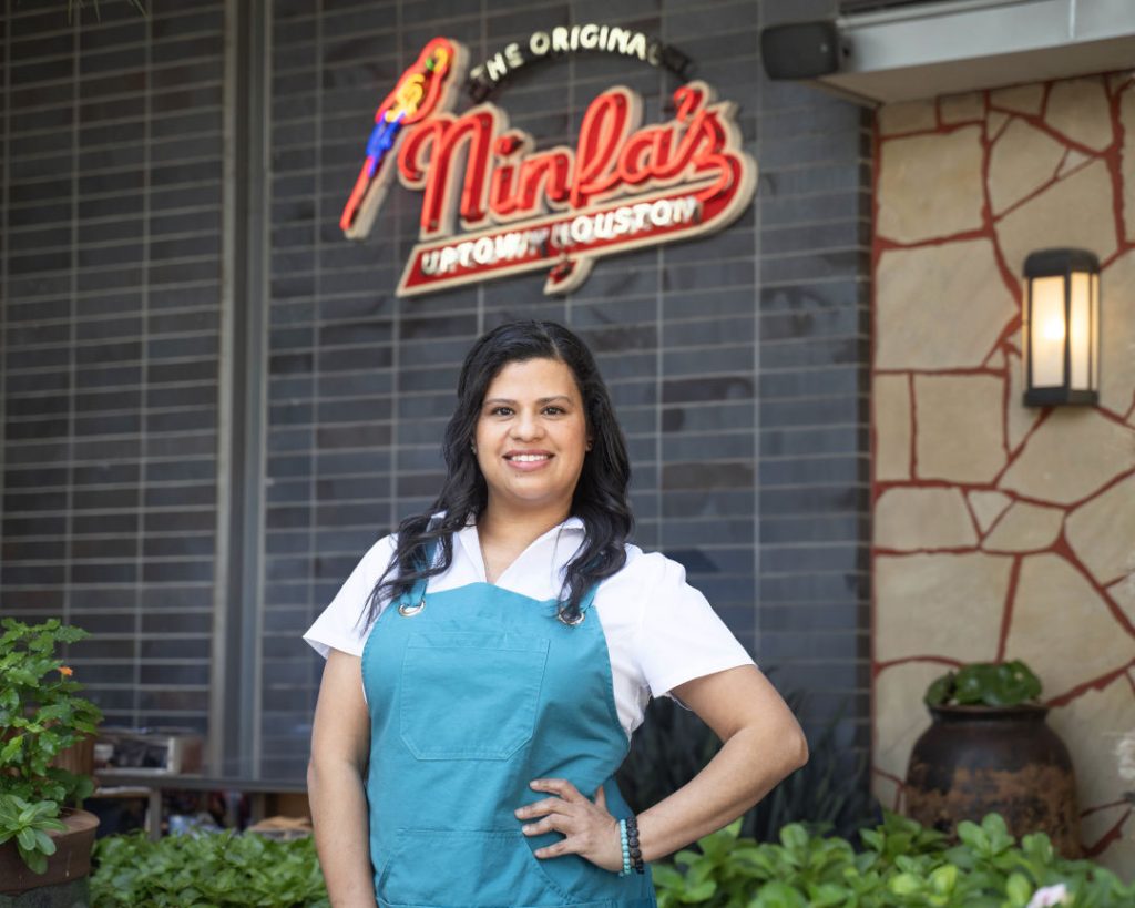 Chef Patricia Delgado stands smiling in front of a Ninfa’s Uptown Houston restaurant sign outdoors.