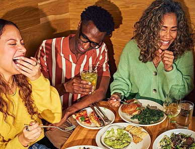 Three friends sitting at a table, laughing and enjoying a meal together with various dishes in front of them.
