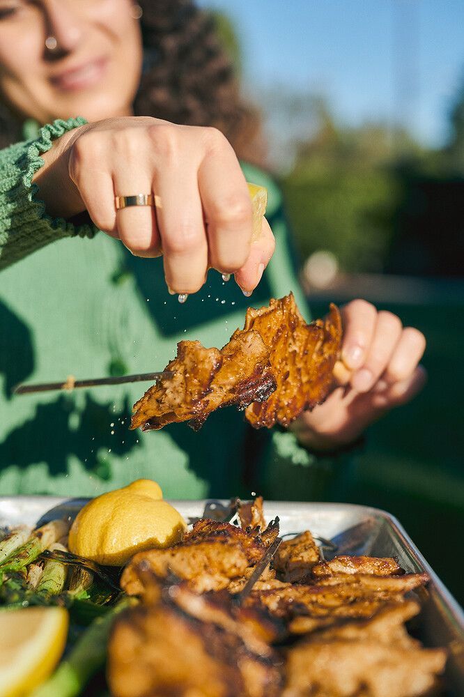 A person squeezes lemon juice over grilled meat outdoors, with more food visible on a tray below.