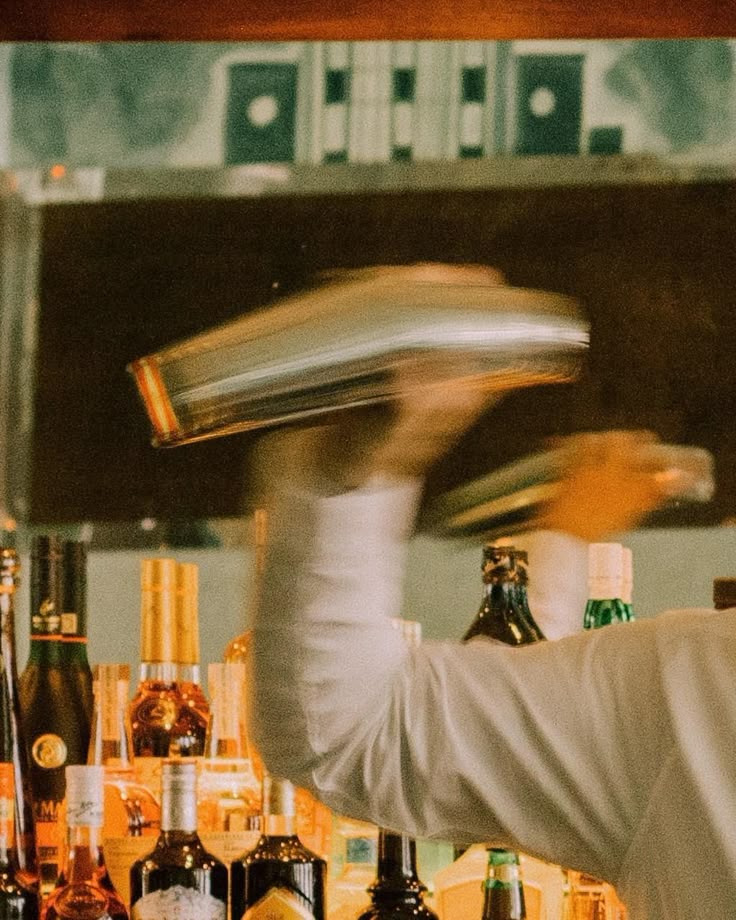 Bartender shaking a cocktail shaker behind a bar with liquor bottles in the background.