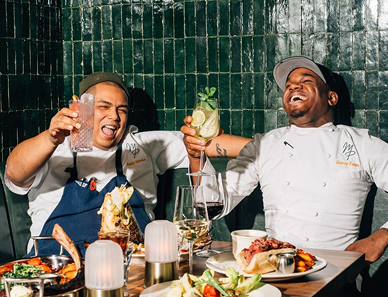 Two chefs in uniform laughing and toasting drinks at a table filled with food in a lively restaurant.