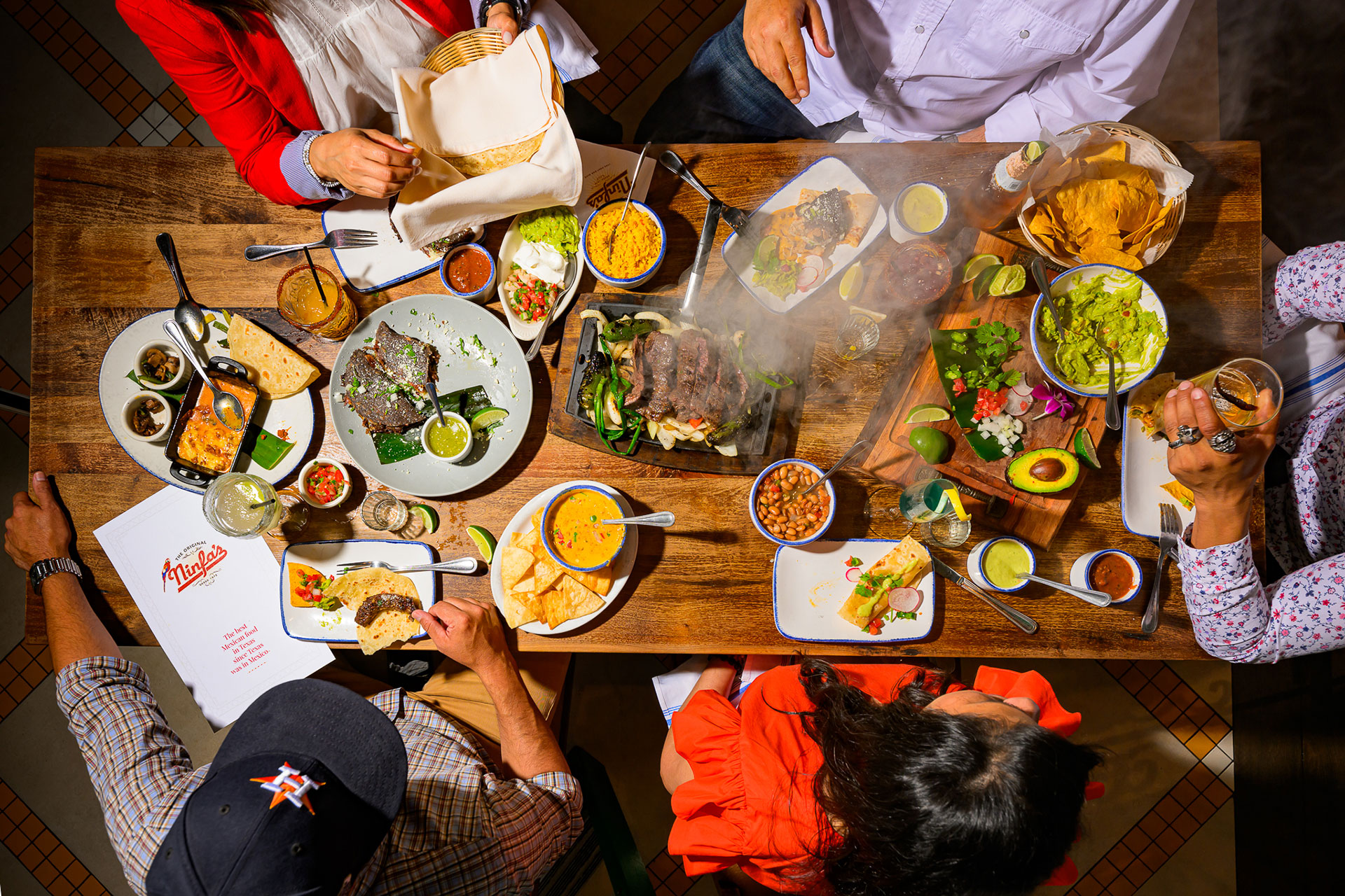 Overhead view of a lively restaurant table at Ninfa's, filled with dishes including sizzling fajitas, tacos, guacamole, rice, beans, chips, salsa, queso, and margaritas, with several people reaching for food and drinks.