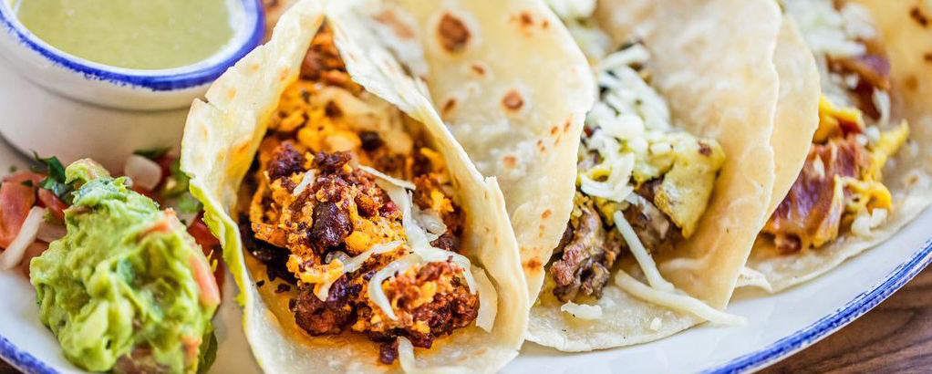 Three Ninfa's breakfast tacos filled with meat and cheese, served with guacamole, salsa, and a cup of green sauce.