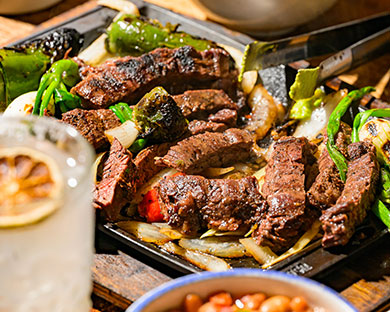 Grilled steak strips with peppers and onions on a hot plate, with a drink and a bowl of beans nearby.