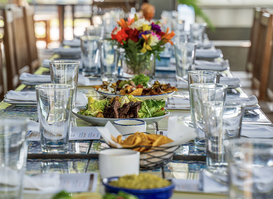 A long dining table set with glasses, plates of food, napkins, and a colorful flower centerpiece in the Front & Side Patio at Ninfa's Uptown.