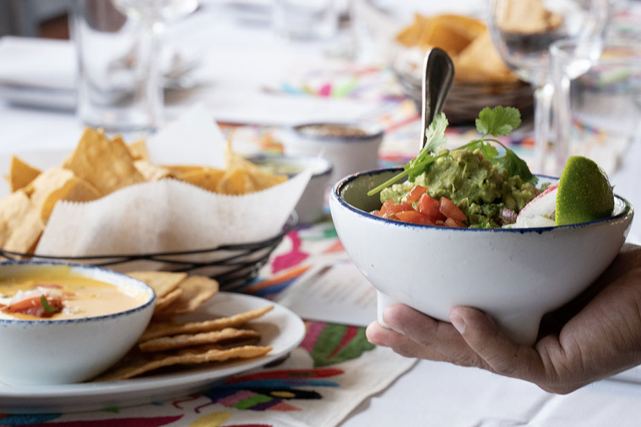 A hand holds a bowl of guacamole with lime; chips and dip are on a colorful table in the background.