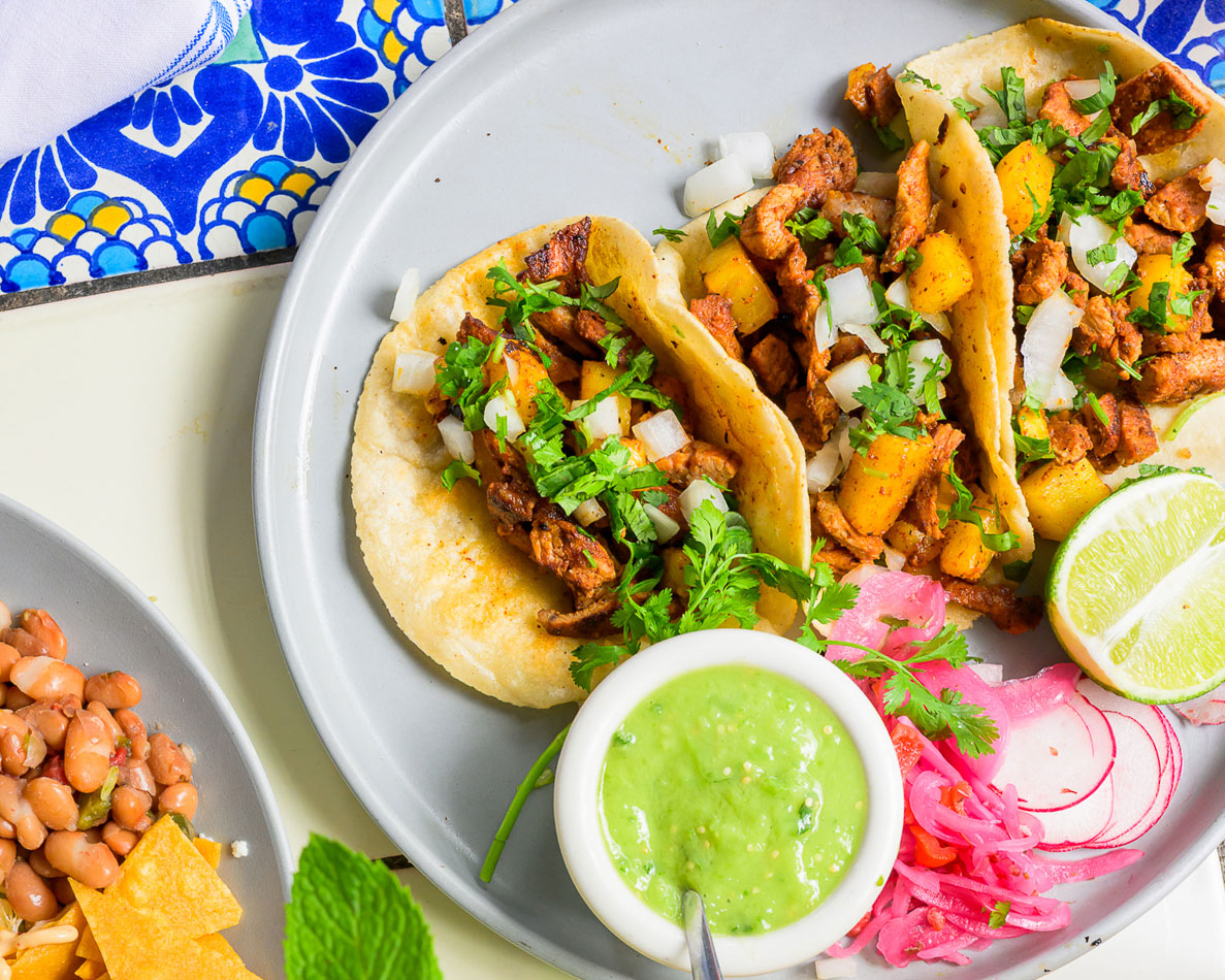 Three tacos with meat, pineapple, onions, and cilantro on a plate with lime, salsa, and pickled onions.