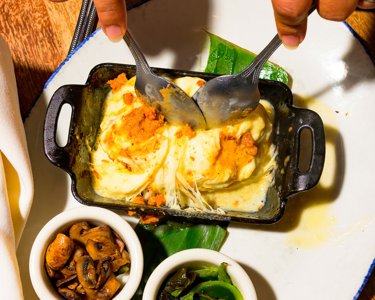 Hands pulling apart stretchy, cheesy chilaquiles in a small black dish, with mushrooms and greens on the side.
