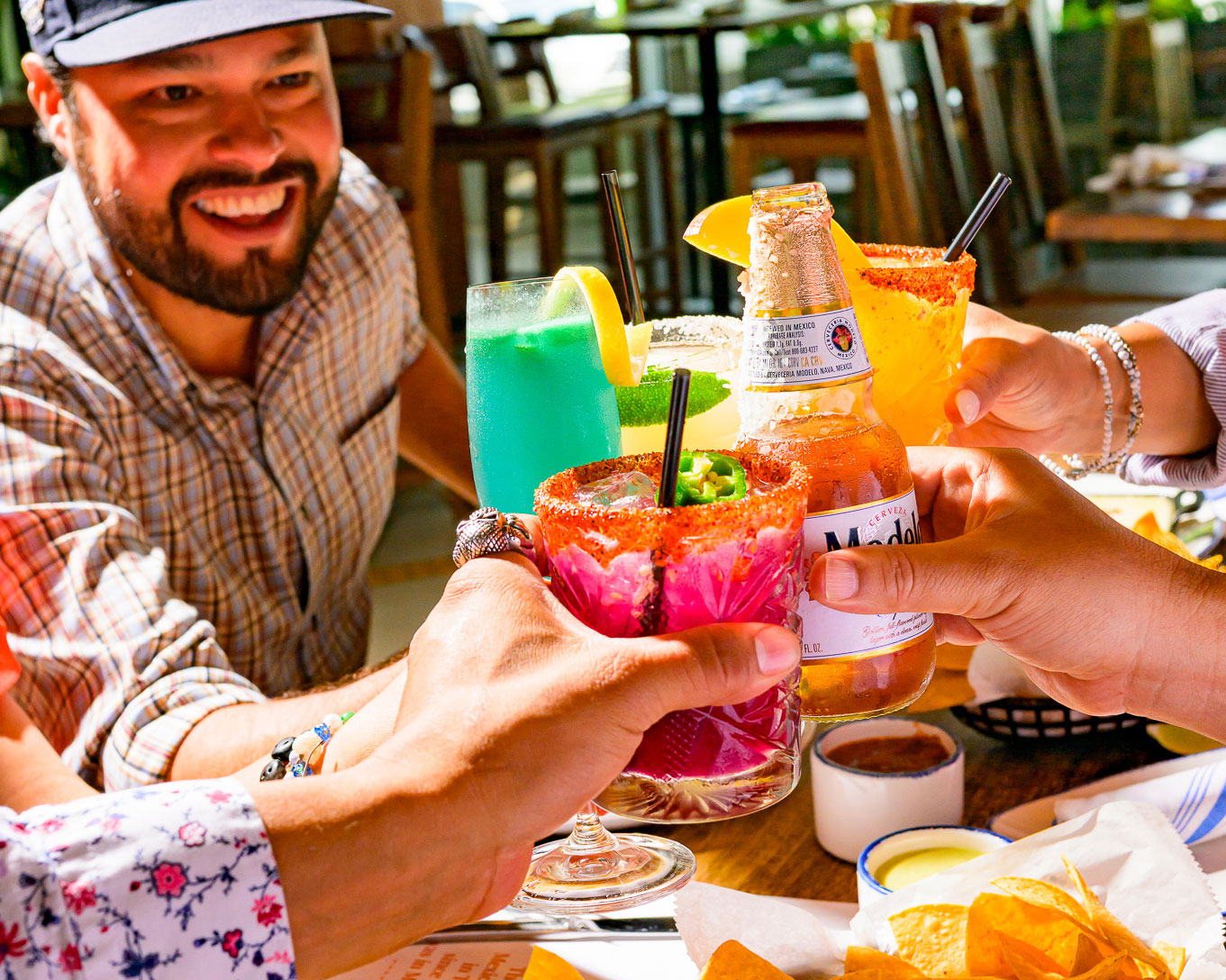 Group of people toasting colorful cocktails and a beer at a sunny table at Ninfa's with chips and dips.