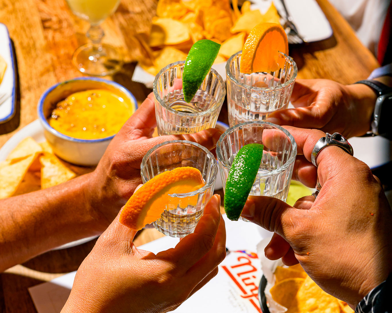 Four hands clink glasses of clear liquor with lime and orange slices, chips and dip on the table.